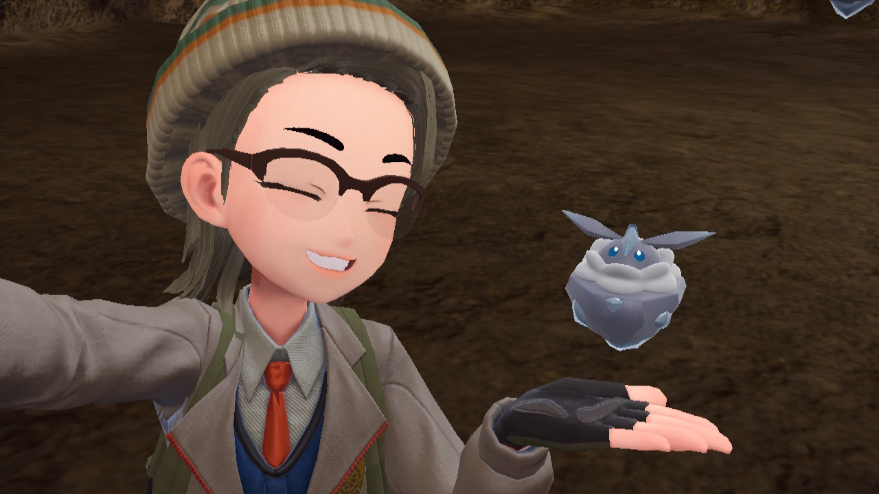 A Pokémon trainer in The Teal Mask DLC smiles and takes a selfie with Carbink, a small blue rock-like Pokémon.