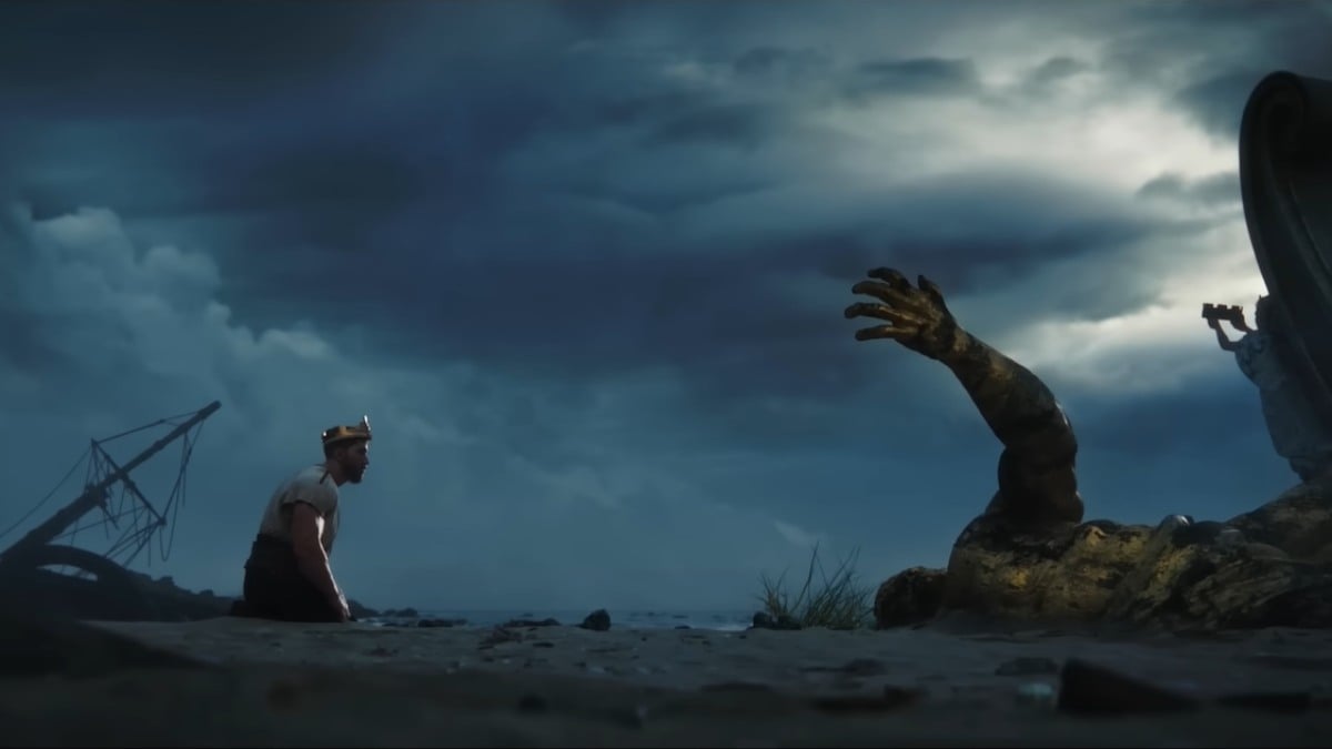 A wide shot of Aelstrom on his knees in the sand, facing his destroyed statue with only an arm sticking out of the ground