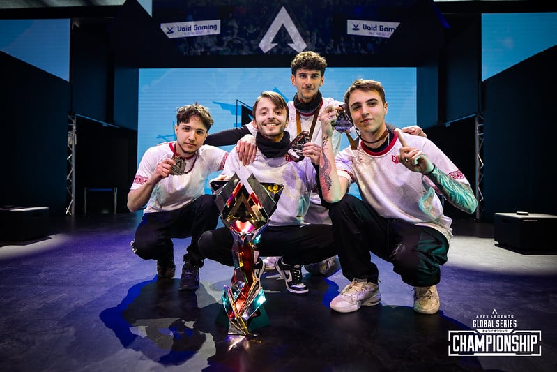 A photograph of GoNext crouching down on stage, holding the Championship trophy
