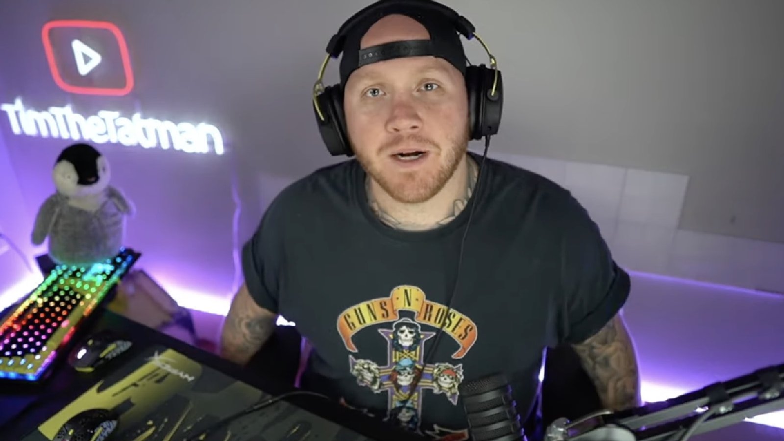 A man in a backwards hat and t-shirt with headphones on talks to the camera.