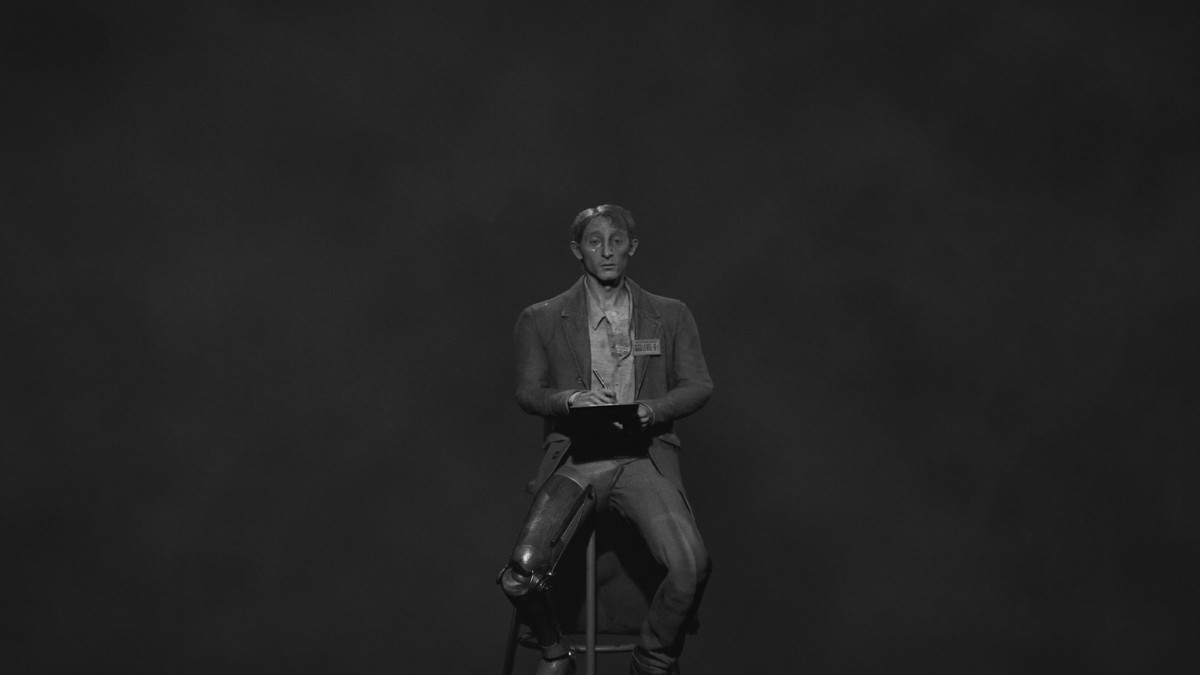 A far away shot of Sean Mehndez sitting on a stool with a clipboard in hand, looking at the camera in black and white shot