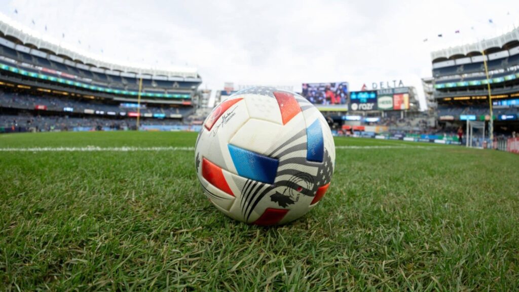 A football placed in the middle of a pitch inside an arena.